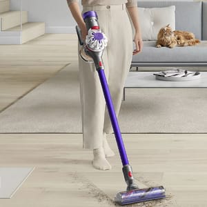 A person is holding a slim, cordless Dyson V8 Animal vacuum with a motorized brush head, designed for pet hair removal, with a cat observing in the background.