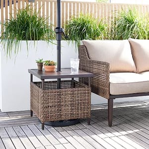 A square-shaped, dark outdoor umbrella table with a central umbrella pole and a wicker armchair next to it on a wooden deck.