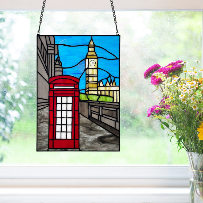 Stained glass window panel featuring a London scene with a red telephone booth and the iconic clock tower. It's hanging in front of a window with a bouquet of colorful flowers nearby.