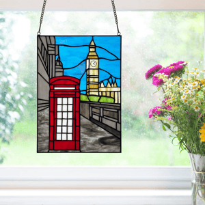 Stained glass window panel featuring a London scene with a red telephone booth and the iconic clock tower. It's hanging in front of a window with a bouquet of colorful flowers nearby.