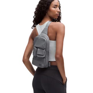 A person wearing a small, gray crossbody bag over one shoulder. The bag has a zippered main compartment and a front pocket with a flap.