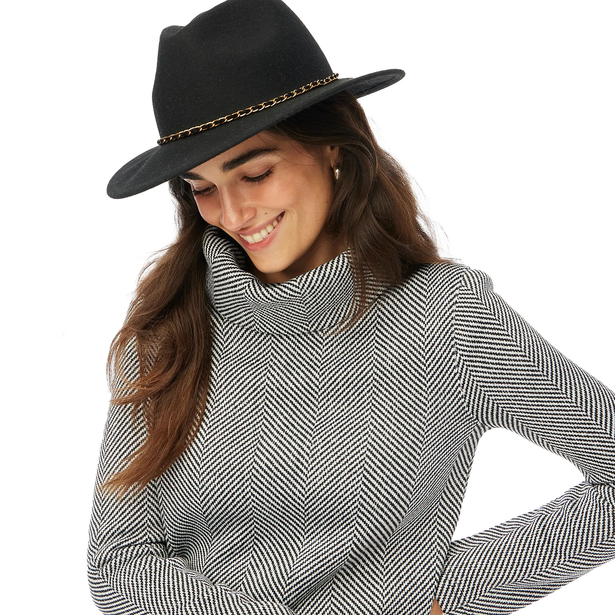 A woman wears a black and white herringbone-patterned funnelneck top, featuring a cozy, high neckline and long sleeves. Her outfit is complemented by a black hat with a chain accent.