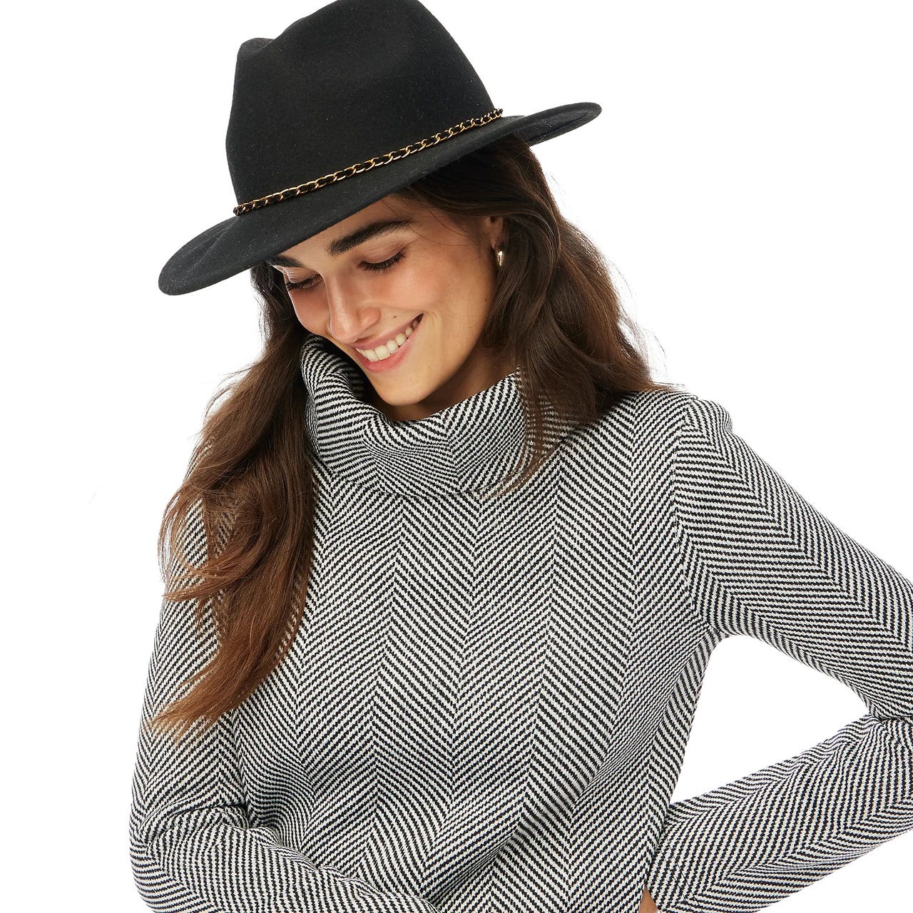 A woman wears a black and white herringbone-patterned funnelneck top, featuring a cozy, high neckline and long sleeves. Her outfit is complemented by a black hat with a chain accent.