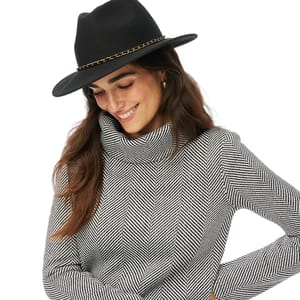 A woman wears a black and white herringbone-patterned funnelneck top, featuring a cozy, high neckline and long sleeves. Her outfit is complemented by a black hat with a chain accent.