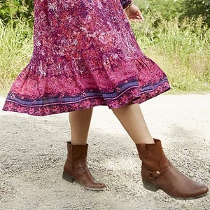 A person is wearing brown ankle boots with buckle detailing and a flowing, pink and blue patterned midi skirt.