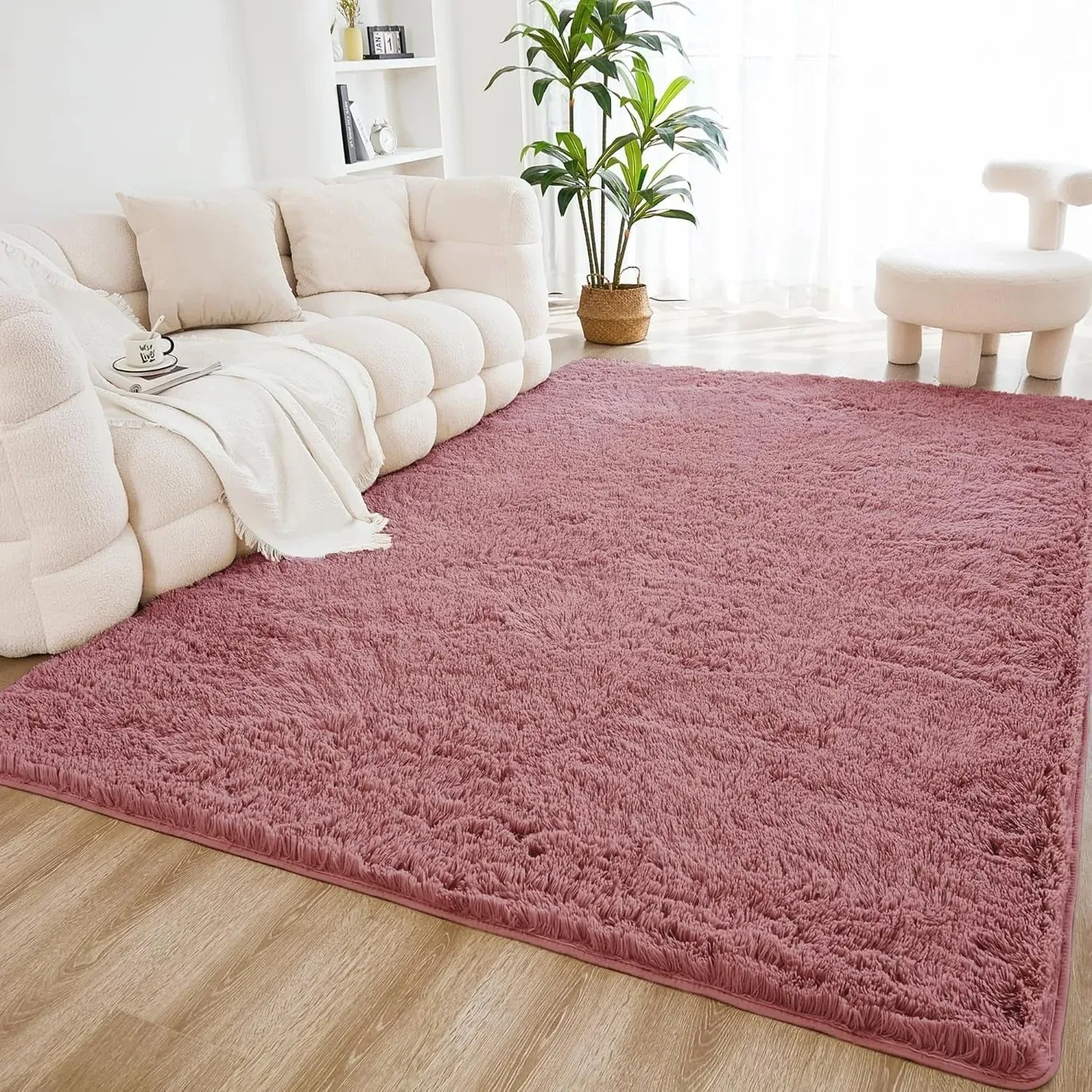A cozy living room setup featuring a fluffy pink rug, a cream-colored sofa with pillows and a blanket, a small white chair, and a potted plant, all set on a wooden floor.