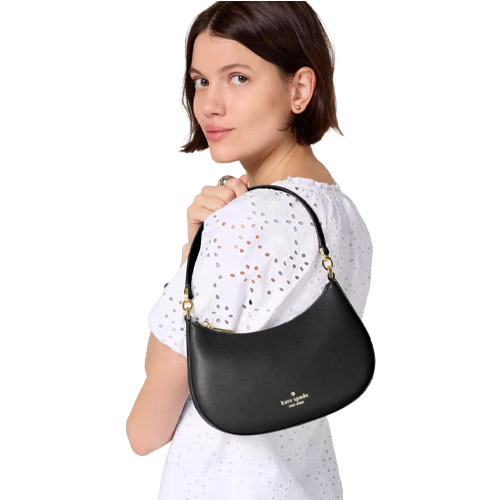 A person wearing a white blouse holds a black Kate Spade handbag with a gold logo, featuring a short strap and a curved design.