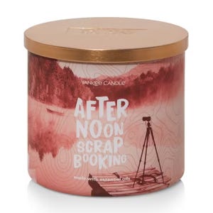Yankee Candle Afternoon Scrapbooking features a design with a serene lakeside scene and a tripod silhouette, packaged in a container with a bronze-colored lid. It is made with essential oils.