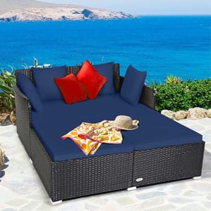 Outdoor wicker patio daybed with dark blue cushions and red throw pillows, set against a coastal backdrop.