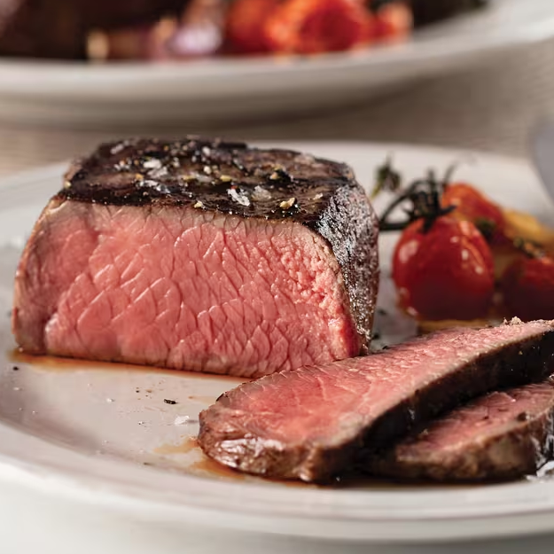 A medium-rare steak is sliced on a plate, garnished with flaky salt. Roasted cherry tomatoes are served alongside.