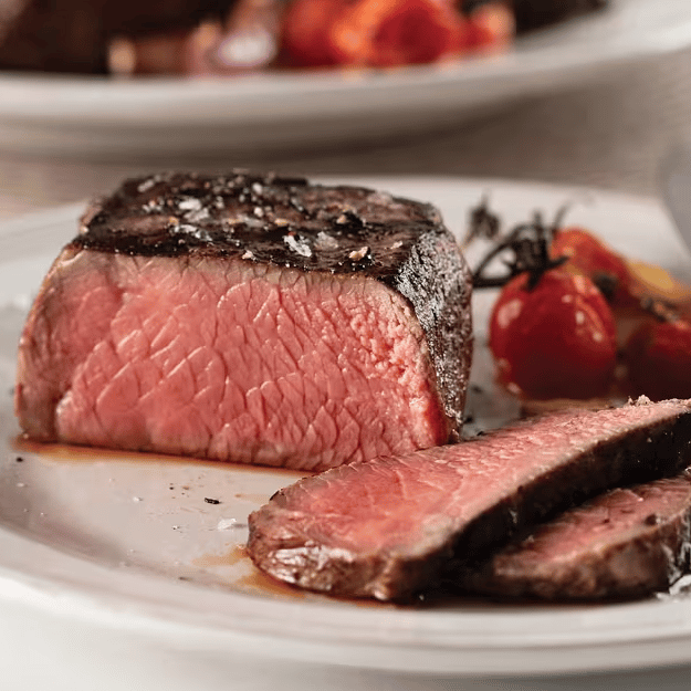 A medium-rare steak is sliced on a plate, garnished with flaky salt. Roasted cherry tomatoes are served alongside.