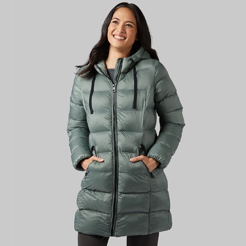 a woman with dark hair wearing a sage green 3/4 length puffer coat against a grey background