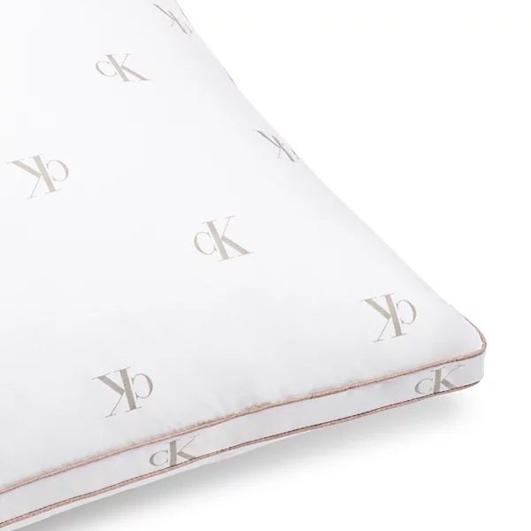 A white pillow featuring the \“cK\“ logo in a repeating pattern with a subtle beige color. The pillow has a piping detail along the edges.