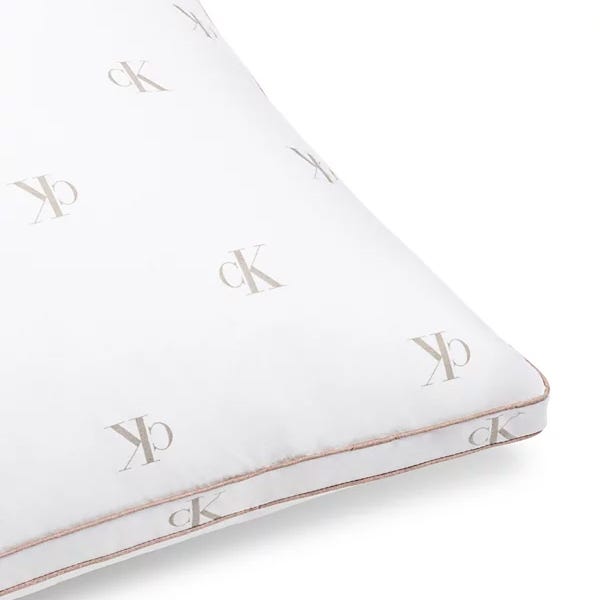 A white pillow featuring the \“cK\“ logo in a repeating pattern with a subtle beige color. The pillow has a piping detail along the edges.