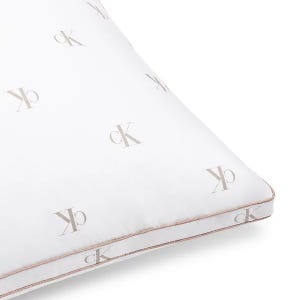 A white pillow featuring the \“cK\“ logo in a repeating pattern with a subtle beige color. The pillow has a piping detail along the edges.