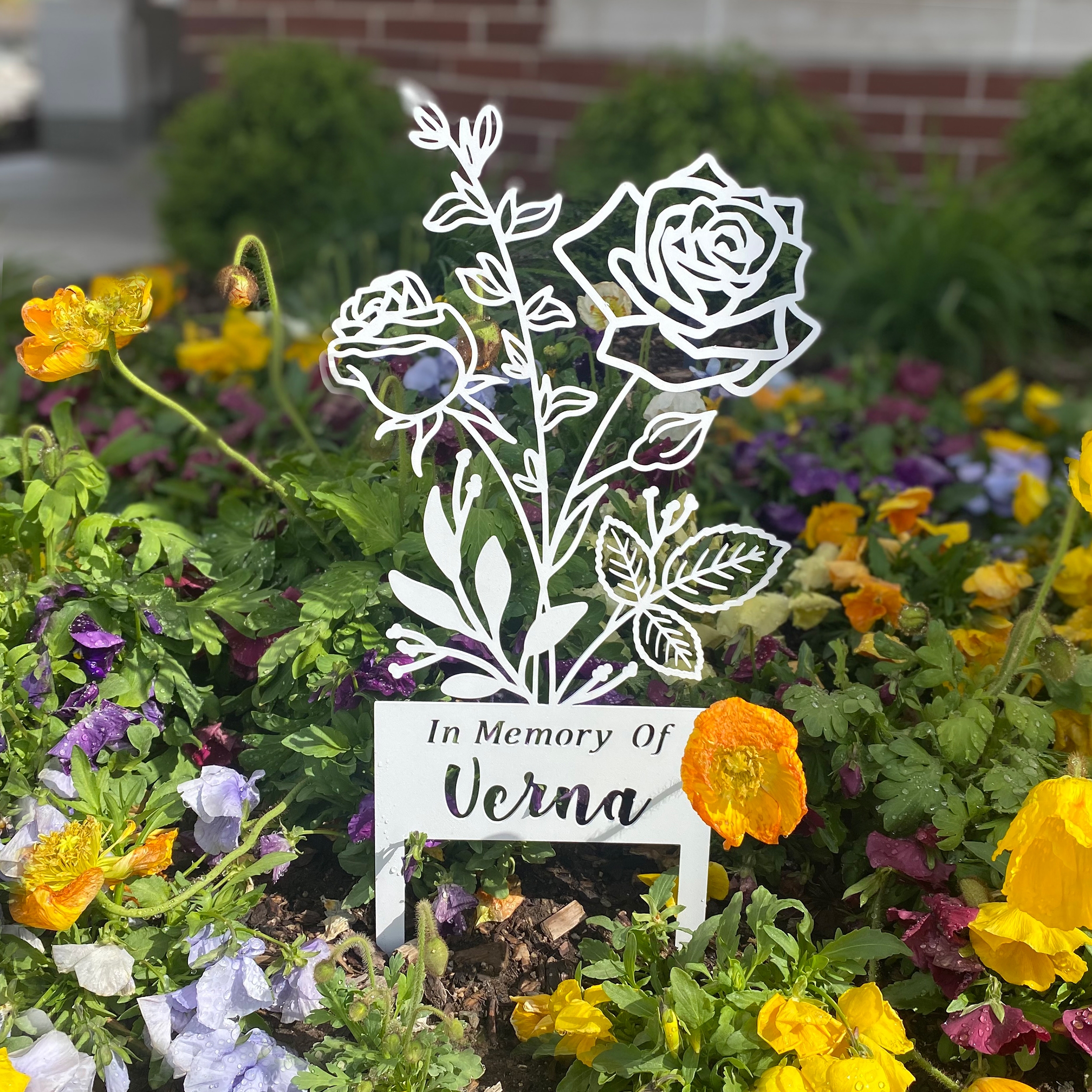 Metal memorial garden marker displaying the text \“In Memory Of Verna,\“ with a rose design, surrounded by colorful flowers.