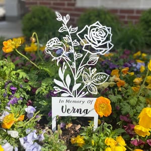 Metal memorial garden marker displaying the text \“In Memory Of Verna,\“ with a rose design, surrounded by colorful flowers.
