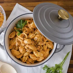 A pot of shell pasta with a creamy sauce, garnished with parsley, sits on a wooden table. The dish is in a gray pot with a lid and brass handle, accompanied by a white napkin and a wooden spoon.