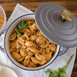 A pot of shell pasta with a creamy sauce, garnished with parsley, sits on a wooden table. The dish is in a gray pot with a lid and brass handle, accompanied by a white napkin and a wooden spoon.