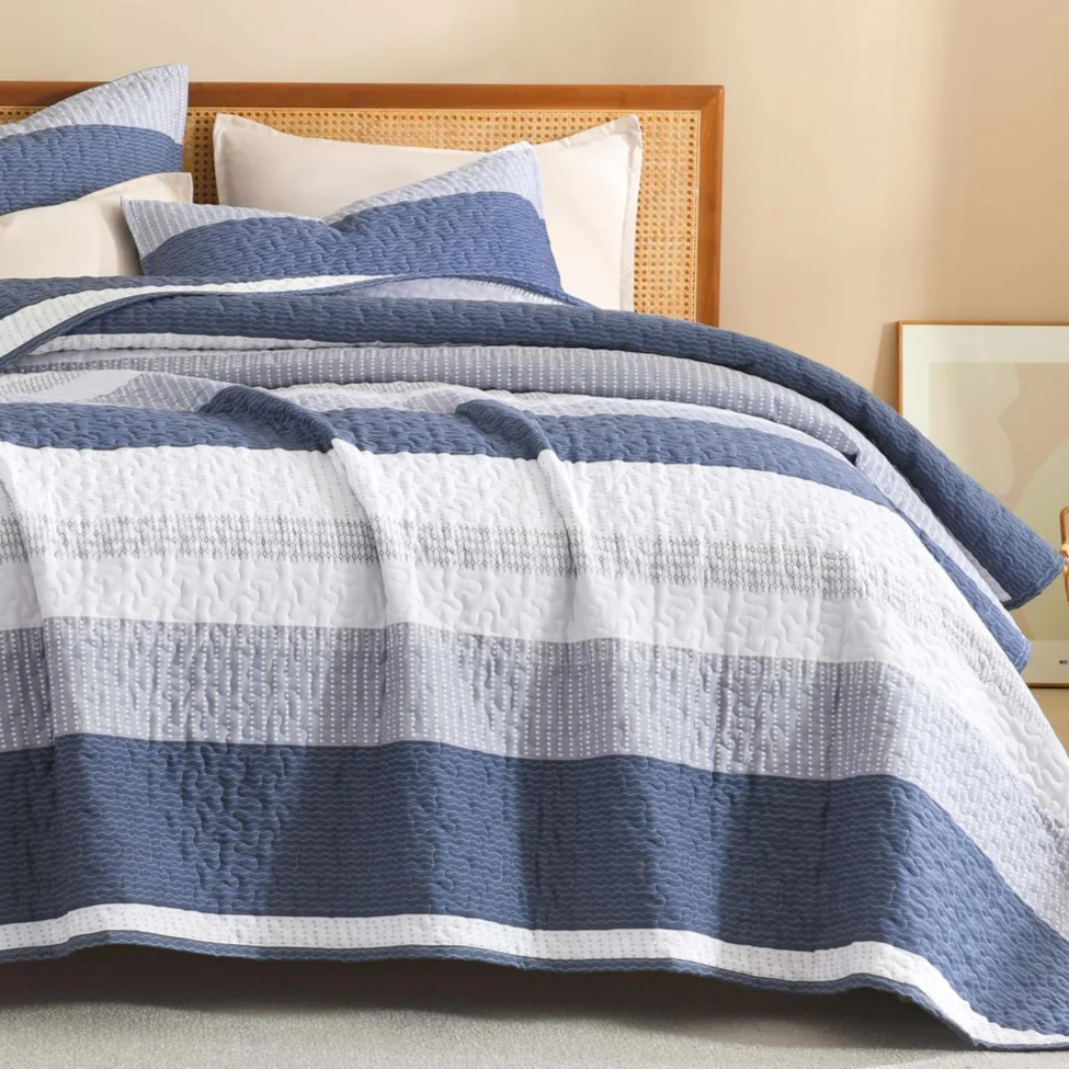 A bed is adorned with a striped quilt and matching pillows in shades of blue, gray, and white, set against a rattan headboard. A framed artwork and woven basket sit nearby.