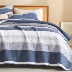 A bed is adorned with a striped quilt and matching pillows in shades of blue, gray, and white, set against a rattan headboard. A framed artwork and woven basket sit nearby.