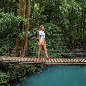 A woman walks on a narrow wooden bridge above a turquoise river, surrounded by lush green jungle. She's wearing a white shirt, tan shorts, and red sneakers.