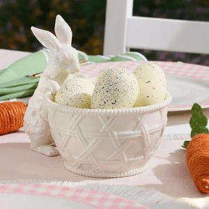 The Lenox Bunny Candy Dish features a white ceramic bowl with a woven pattern, accompanied by a ceramic rabbit figure. It holds speckled eggs and is placed on a pink gingham tablecloth next to a decorative carrot.