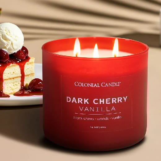 A lit Colonial Candle labeled \“Dark Cherry Vanilla,\“ with scents of black cherry, woods, and vanilla, is featured beside a slice of cherry-topped cake with ice cream.