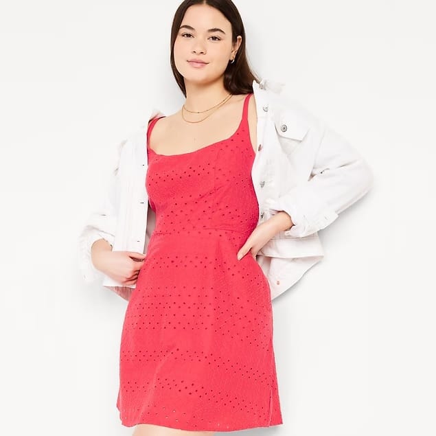 A woman is wearing a red sleeveless eyelet dress and a white denim jacket, accessorized with a simple necklace.
