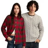 A woman in a red and black plaid shirt stands next to a man wearing a light gray cable-knit sweater. Both are dressed casually.