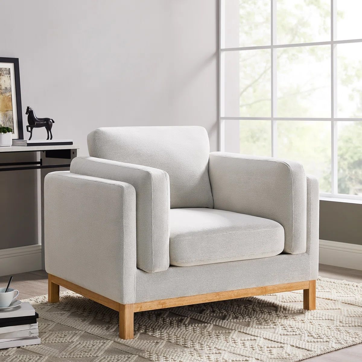 Armchair with light gray upholstery, wooden legs, and a contemporary design, placed on a textured rug in a well-lit room with large windows.