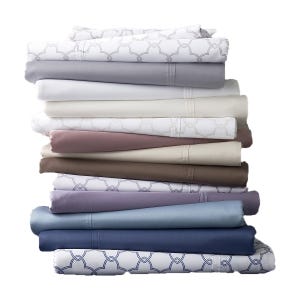 A stack of folded bed sheets in various colors and patterns, including white, beige, brown, blue, lavender, and ones with geometric designs.