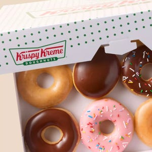 A Krispy Kreme doughnut box partially opened, revealing a variety of doughnuts: glazed, chocolate-covered, one with pink icing and sprinkles, and chocolate with sprinkles. The box features the signature green polka dot design and Krispy Kreme logo.