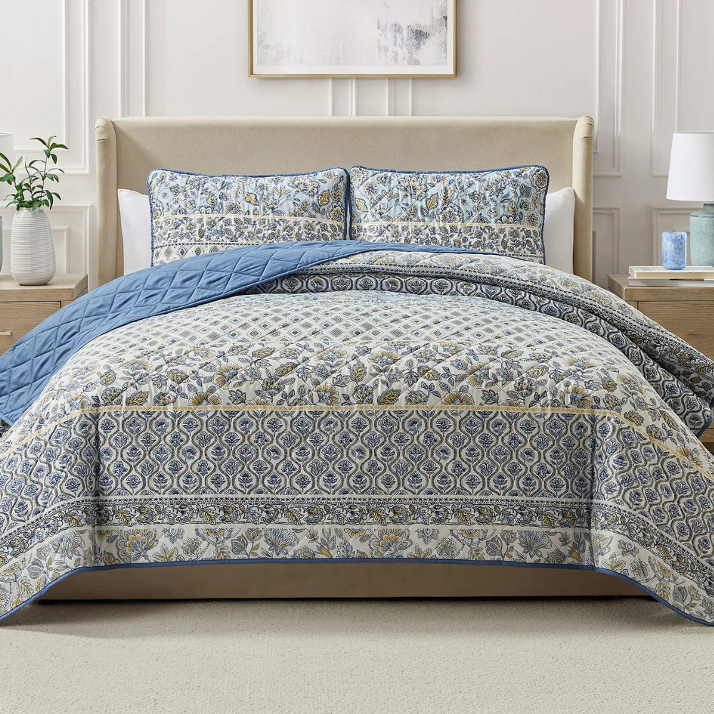 A bed is adorned with a patterned quilt and matching pillow shams in blue and beige hues, featuring floral and geometric designs, complemented by a plain blue underside.