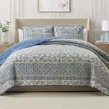 A bed is adorned with a patterned quilt and matching pillow shams in blue and beige hues, featuring floral and geometric designs, complemented by a plain blue underside.