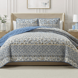 A bed is adorned with a patterned quilt and matching pillow shams in blue and beige hues, featuring floral and geometric designs, complemented by a plain blue underside.
