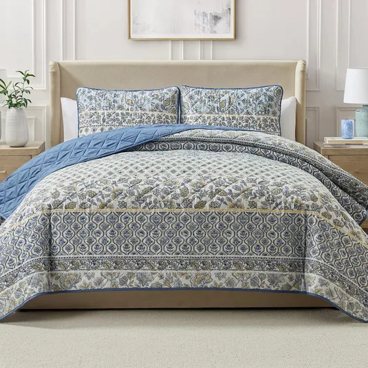 A bed is adorned with a patterned quilt and matching pillow shams in blue and beige hues, featuring floral and geometric designs, complemented by a plain blue underside.