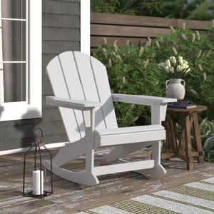 A white Adirondack-style rocking chair is on a deck next to a wooden side table with a vase of flowers. A metal lantern with a candle is positioned on the floor beside the chair.