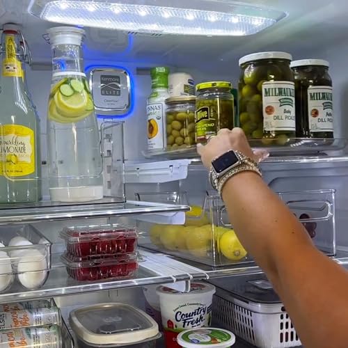 A person's hand is reaching for a container on a clear, turntable-style shelf within a refrigerator stocked with items like lemonade, olives, and lemons.