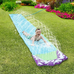 A child is sliding on a water slide positioned on grass with water sprinkling from the sides.