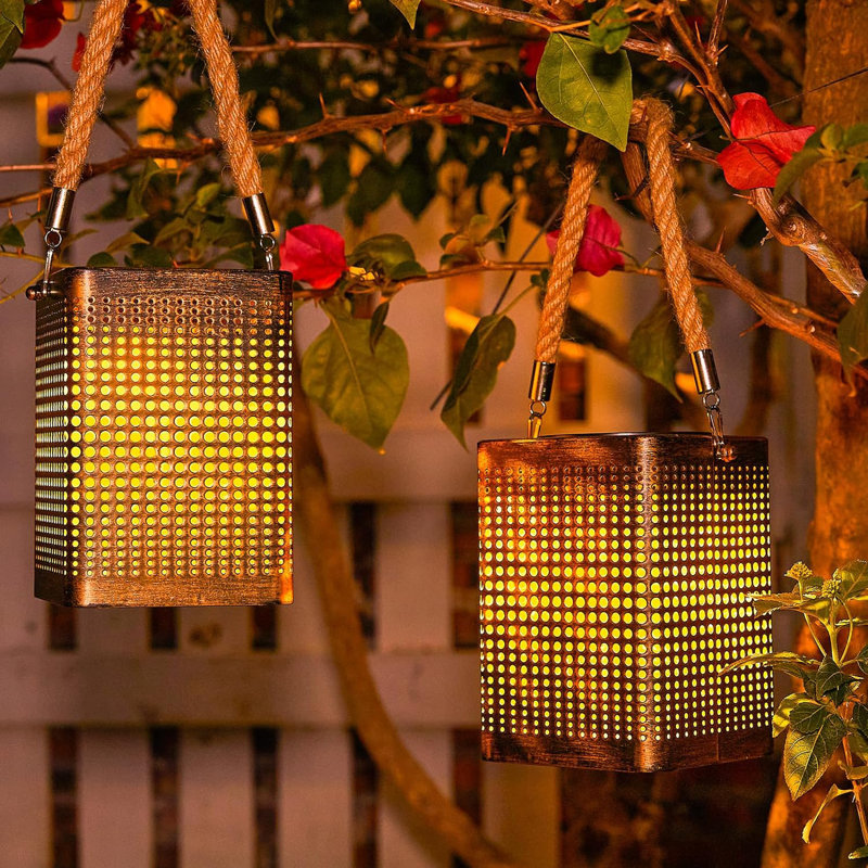 Two cylindrical outdoor lanterns with warm LED lights are hanging from tree branches by ropes.