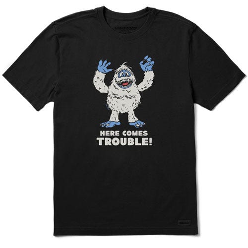 Black T-shirt featuring a cartoon yeti with blue hands and feet, and the text \“HERE COMES TROUBLE!\“ written below the image.