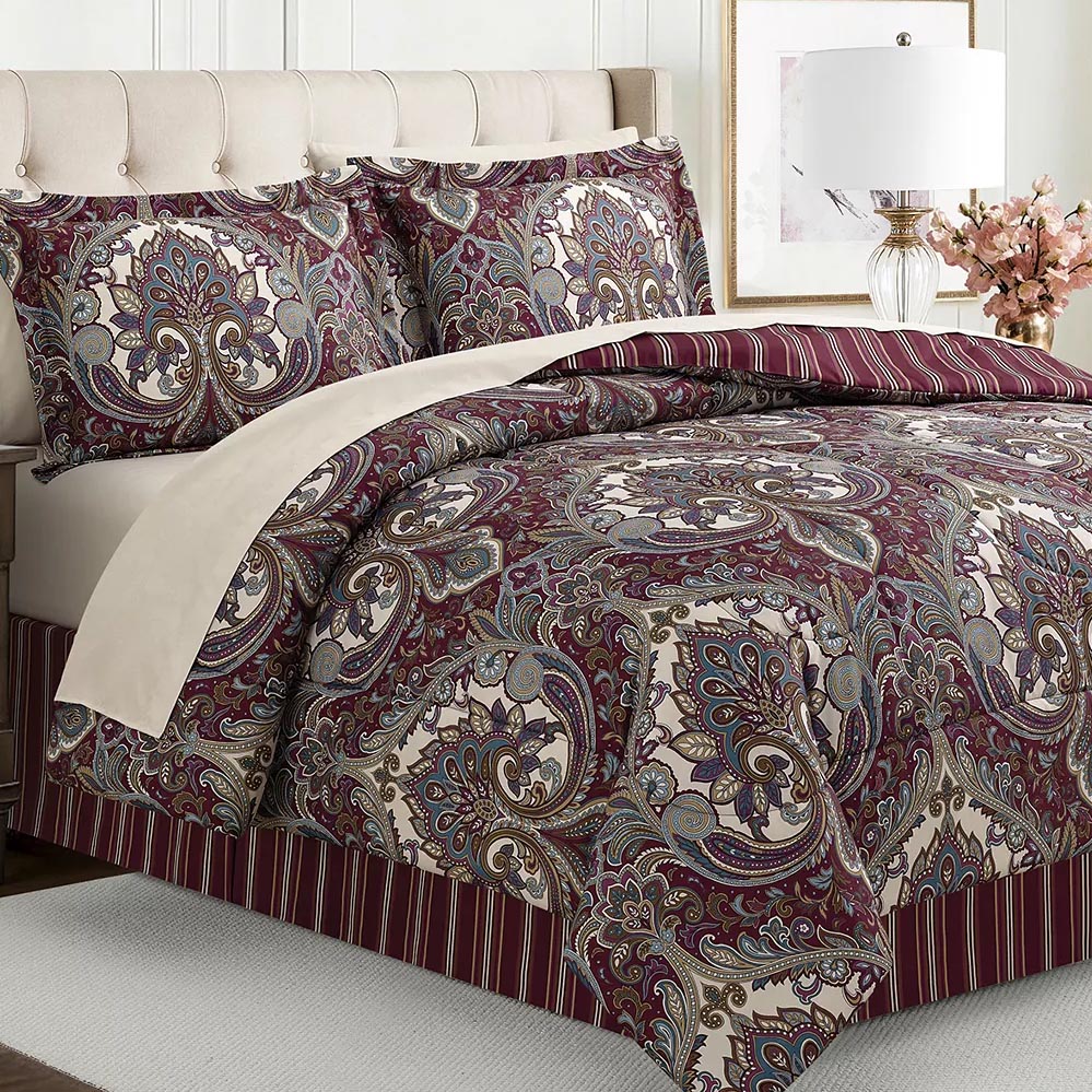 A bed is shown with a paisley pattern comforter set in burgundy, cream, and blue tones, accompanied by matching pillow shams. The bed features a striped bed skirt and is flanked by a beige upholstered headboard, a side lamp, and decorative flowers.