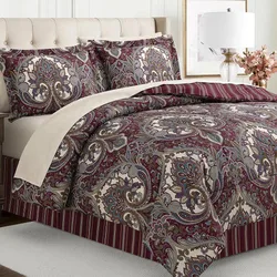 A bed is shown with a paisley pattern comforter set in burgundy, cream, and blue tones, accompanied by matching pillow shams. The bed features a striped bed skirt and is flanked by a beige upholstered headboard, a side lamp, and decorative flowers.
