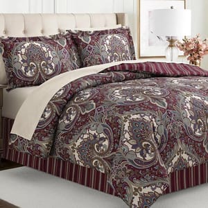A bed is shown with a paisley pattern comforter set in burgundy, cream, and blue tones, accompanied by matching pillow shams. The bed features a striped bed skirt and is flanked by a beige upholstered headboard, a side lamp, and decorative flowers.