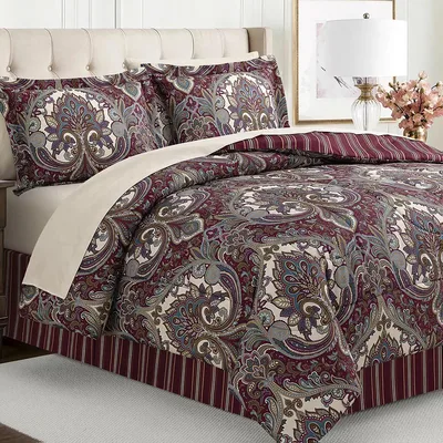 A bed is shown with a paisley pattern comforter set in burgundy, cream, and blue tones, accompanied by matching pillow shams. The bed features a striped bed skirt and is flanked by a beige upholstered headboard, a side lamp, and decorative flowers.