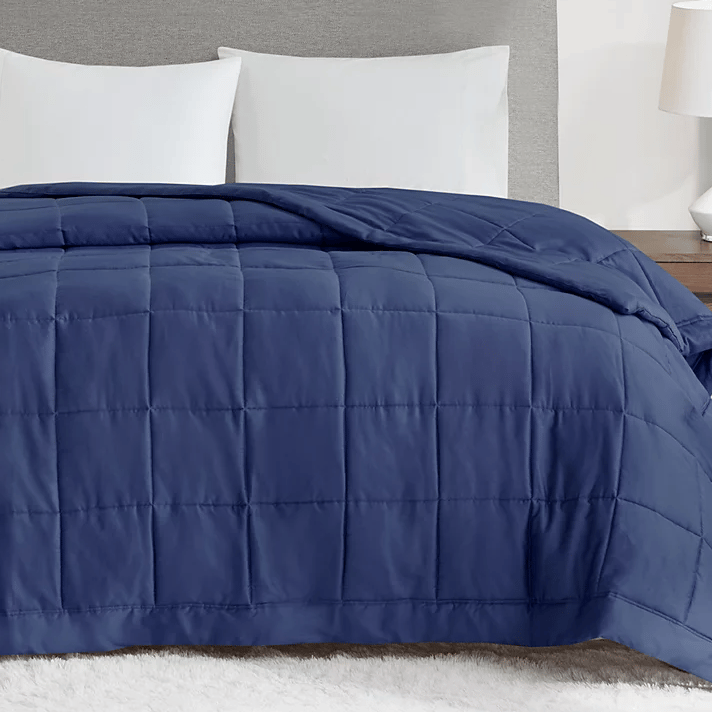 A navy blue, quilted, down-alternative blanket is displayed on a bed. It features a smooth surface with square stitching and is accompanied by white pillows against a neutral headboard.