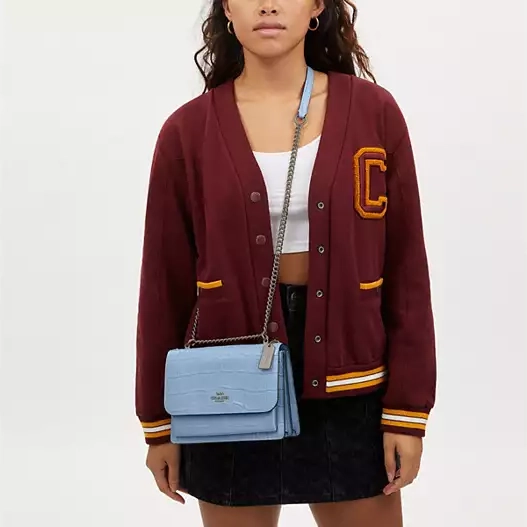 A woman wearing a varsity jacket, a crop top, a dark mini skirt, and carrying a light blue shoulder bag.