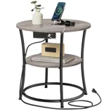A round side table with black metal legs features a wooden top, two shelves, built-in power outlets and USB ports, and is shown with a charging phone and smartwatch, a small vase, books, and an alarm clock.