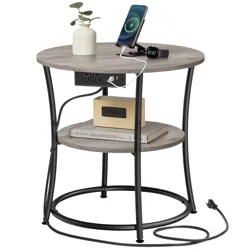 A round side table with black metal legs features a wooden top, two shelves, built-in power outlets and USB ports, and is shown with a charging phone and smartwatch, a small vase, books, and an alarm clock.